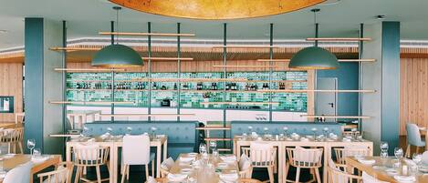A restaurant with wooden tables and chairs, a bar area with shelves of bottles, and a large circular ceiling light.