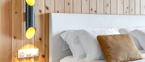 A bedroom with a wooden wall panel, a bedside table with a lamp, a bed with white bedding and pillows, and a power outlet on the wall.