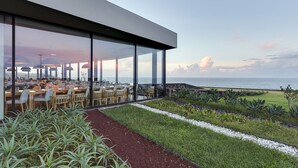 A modern outdoor dining area with glass walls, a grassy garden, and a view of the sea. - Pedras do Mar Resort & SPA (Ponta Delgada)