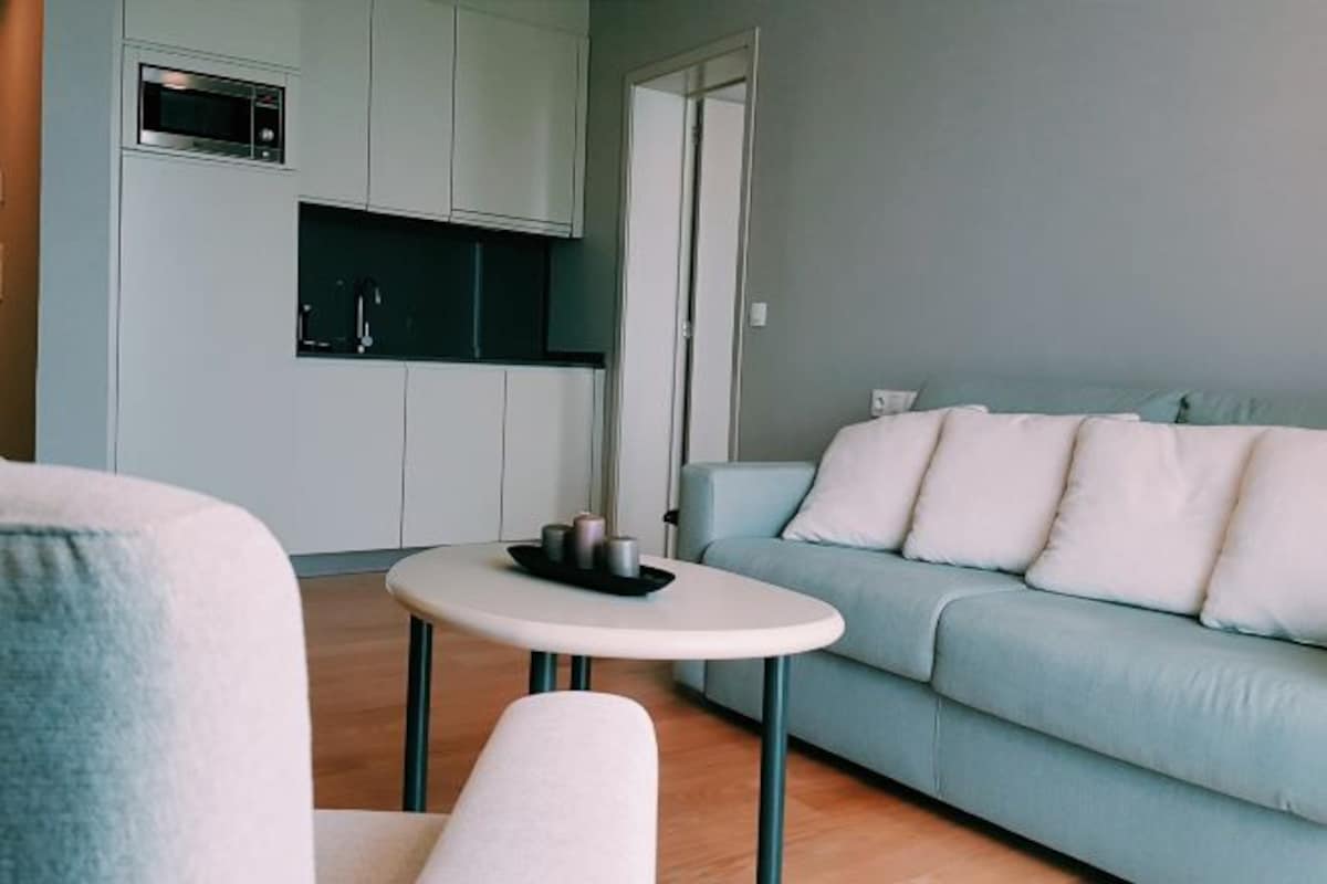 A modern living room with a light blue sofa, a small round table with a tray, and a kitchen area with white cabinets and a microwave.