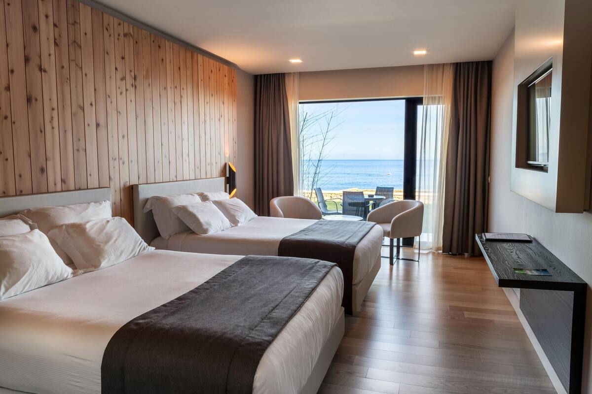 A hotel room with two beds, a desk, a chair, and a view of the sea.