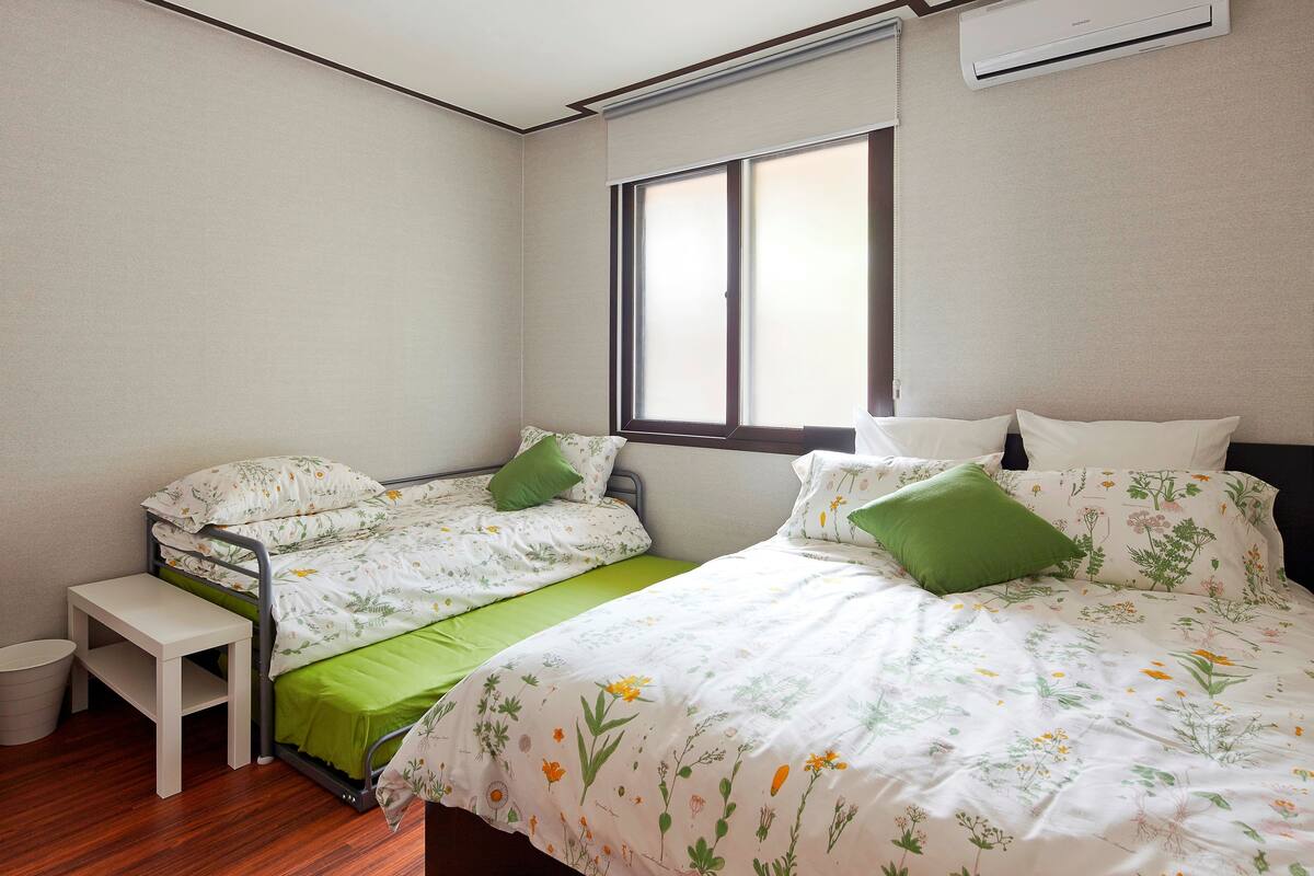 Een slaapkamer met twee bedden, een klein wit bankje en een raam met jaloezieën.