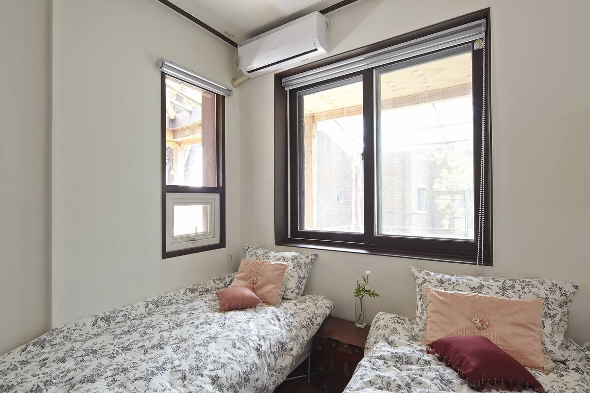Een slaapkamer met twee eenpersoonsbedden, een klein nachtkastje met een vaas en een raam met zonwering.