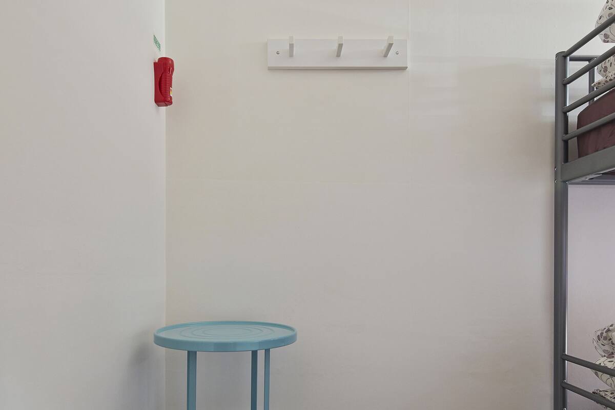 Een hoekzaal met een blauwe drielaagse tafel, een blauwgroene prullenbak, een wit inbouw lichtschakelaar en een metalen stapelbed.