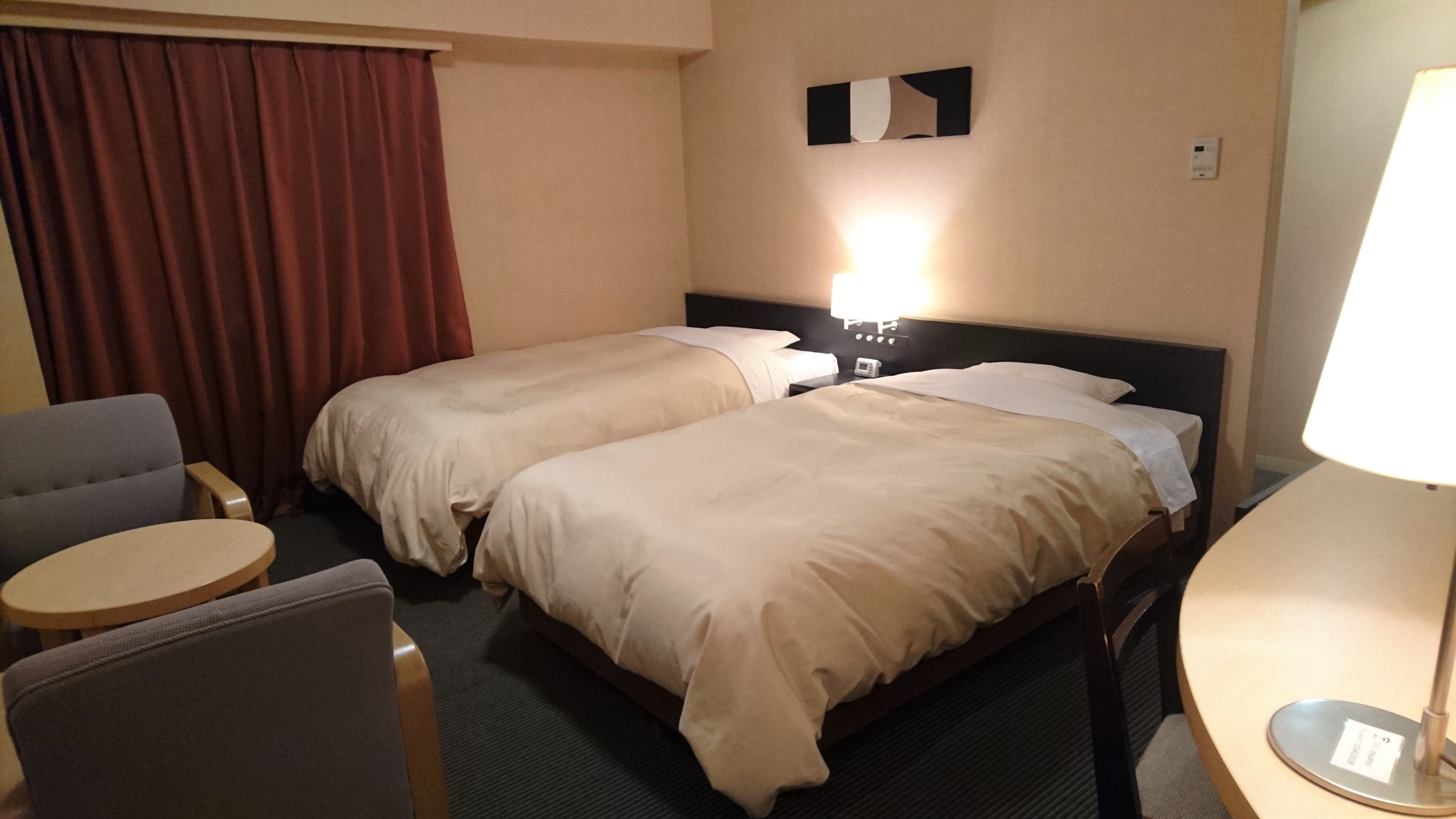 A hotel room with two beds, a small table, a chair, and a lamp.