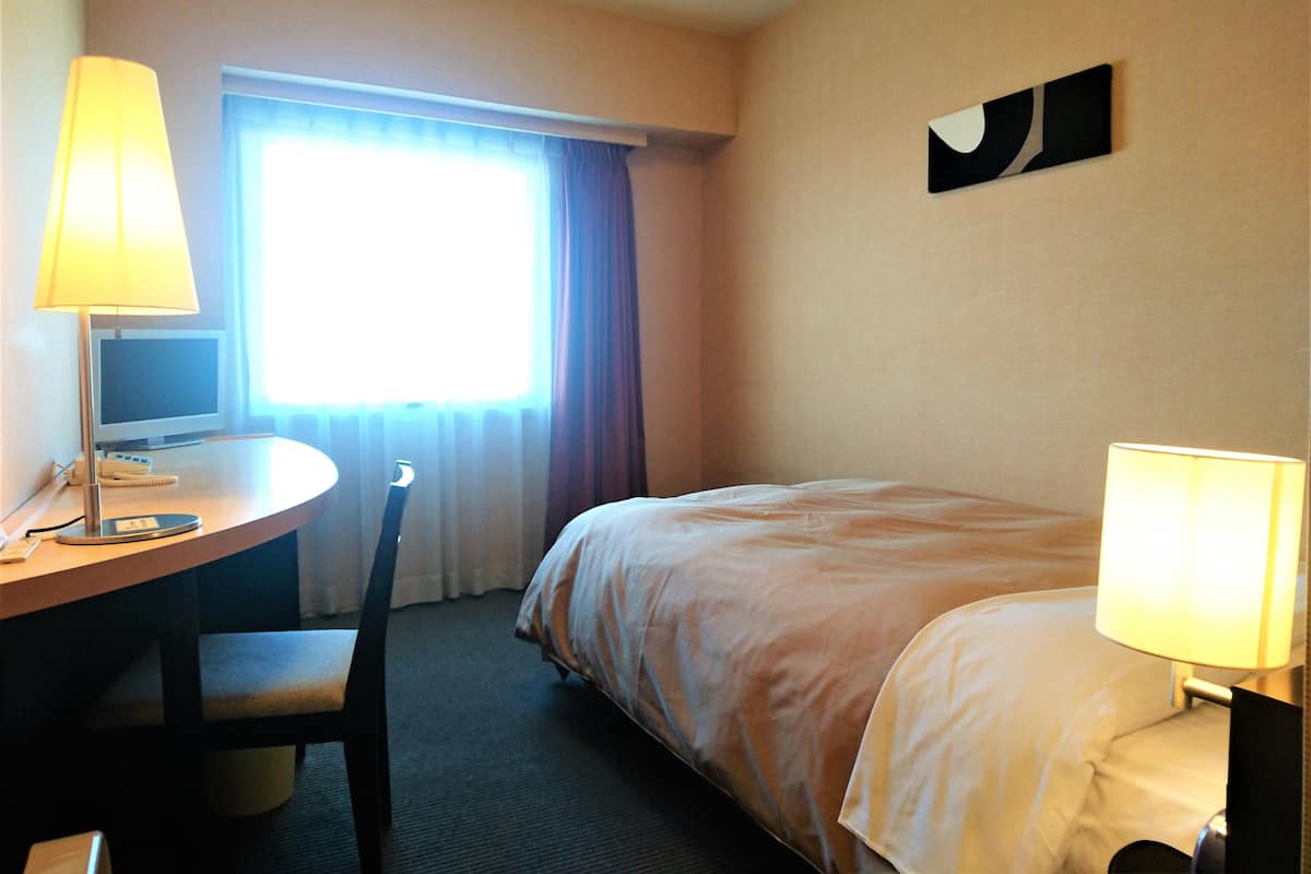 Una habitació d'hotel amb llit, escriptori amb làmpada, cadira i finestra amb cortines.