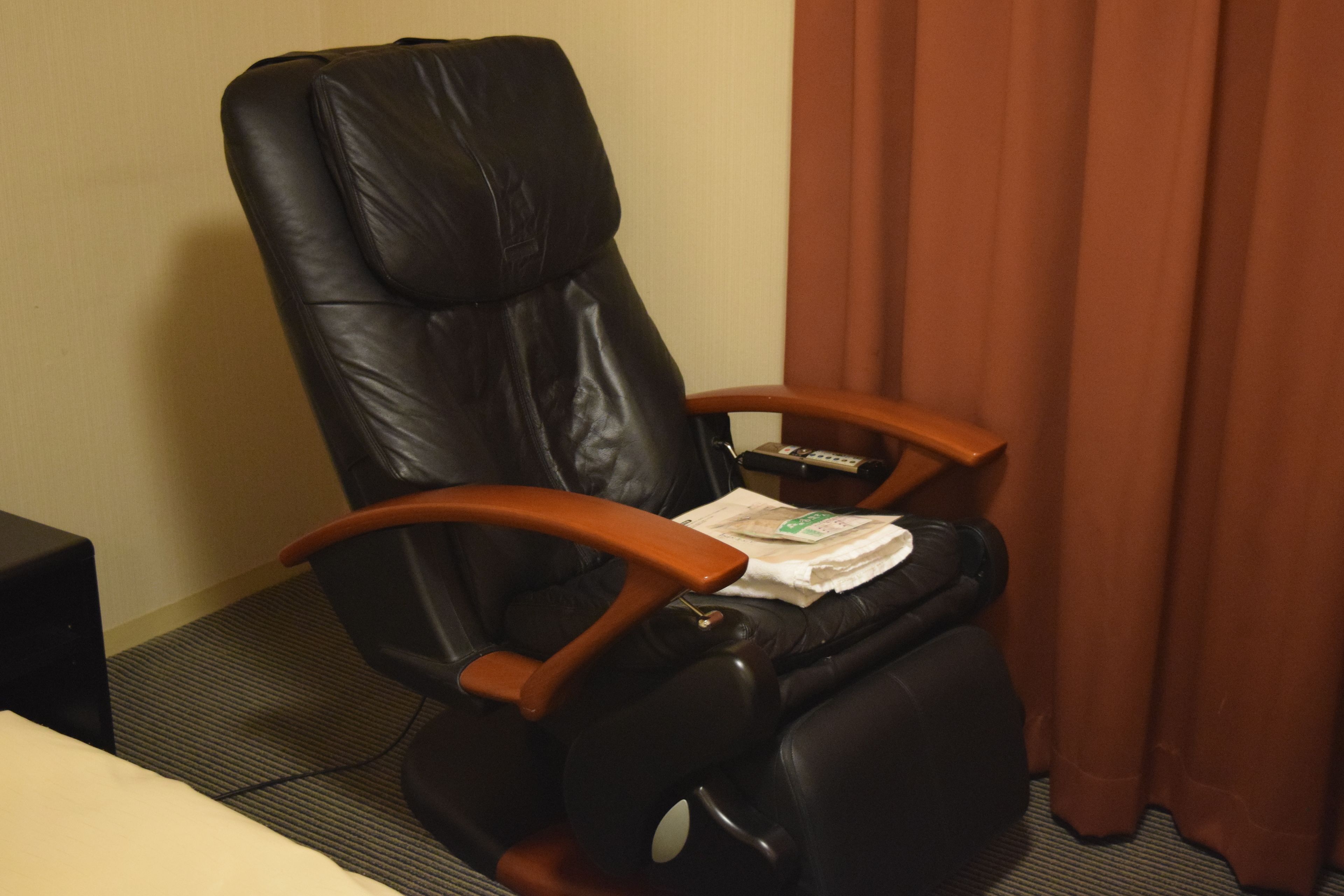 Una silla de masaje de cuero negro con reposabrazos de madera y reposapiés, ubicada en una habitación con cortina marrón y suelo alfombrado.
