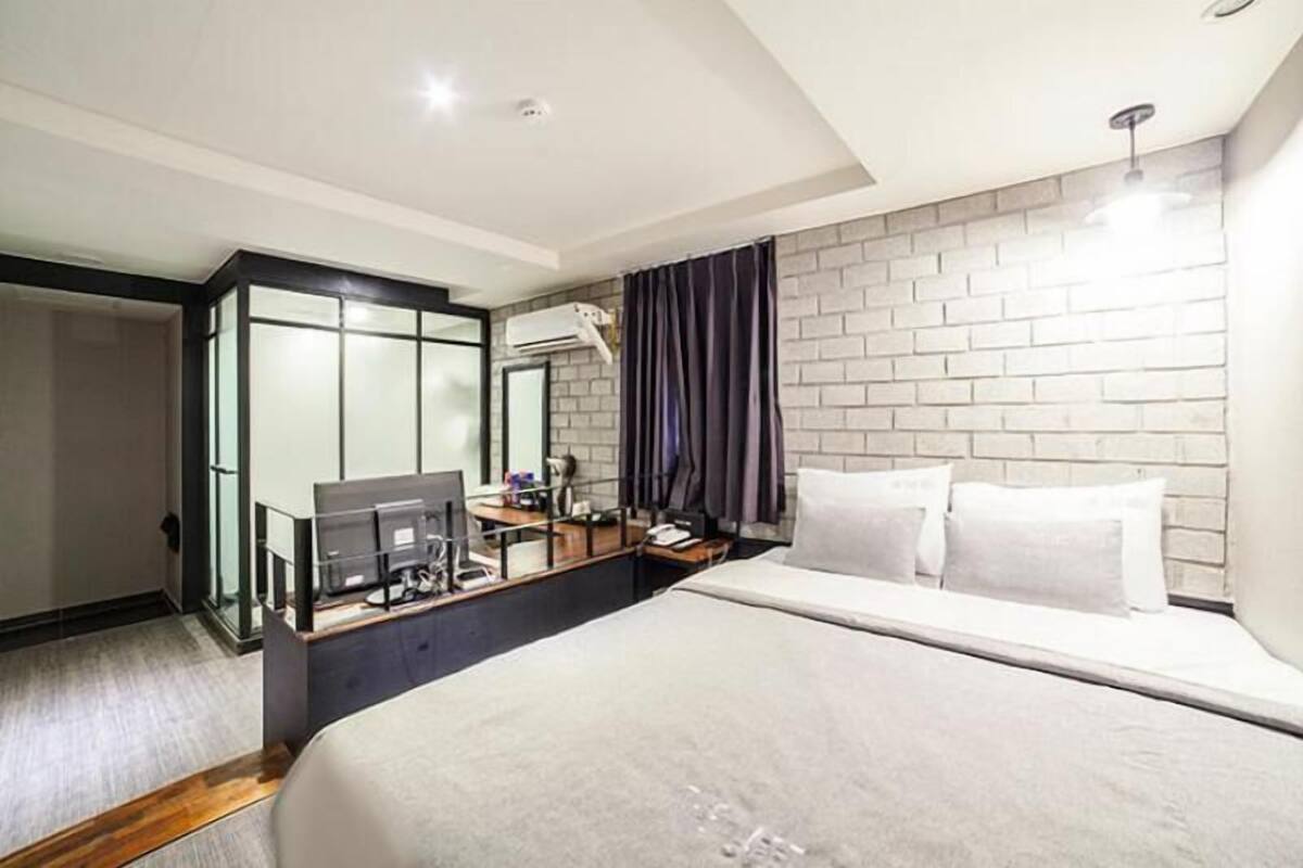 A modern hotel room with a large bed, a desk with a computer, a mirror, and a brick-patterned wall.