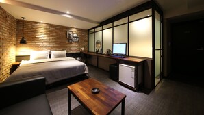 Quarto de hotel moderno com parede de destaque em tijolo, cama bem arrumada, pequena mesa de escritório com computador e cozinha compacta.