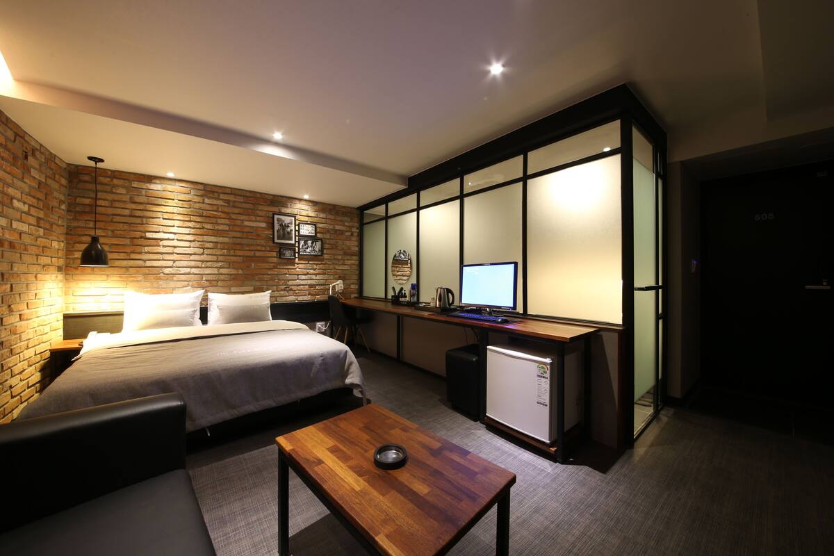 A modern hotel room with a brick accent wall, a neatly made bed, a small desk with a computer, and a compact kitchenette.