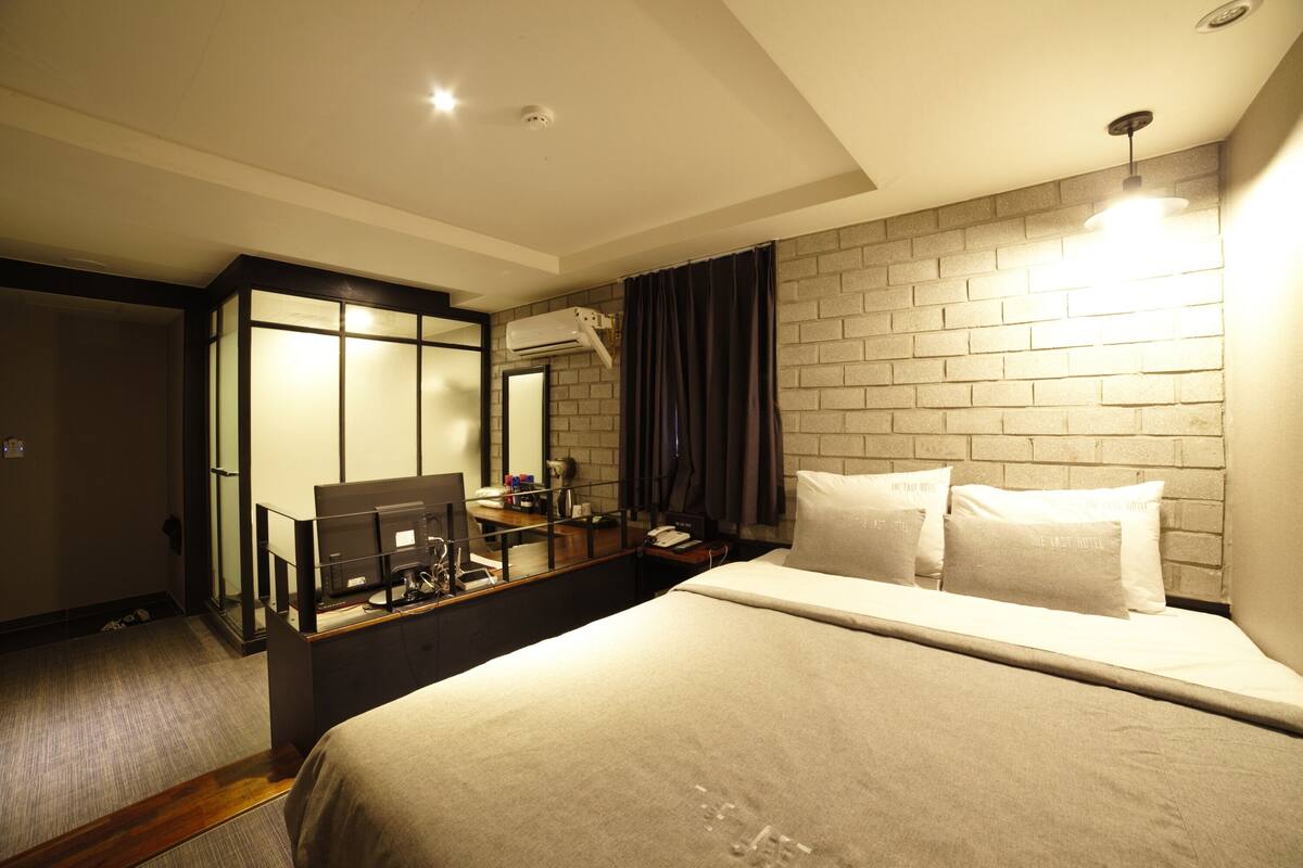 A hotel room with a bed, a desk with a computer, a mirror, and a brick wall.