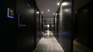 Um corredor comprido e estreito, com design moderno, apresentando um botão de emergência digital, uma porta com teclado numérico e uma placa indicando a sala 505.