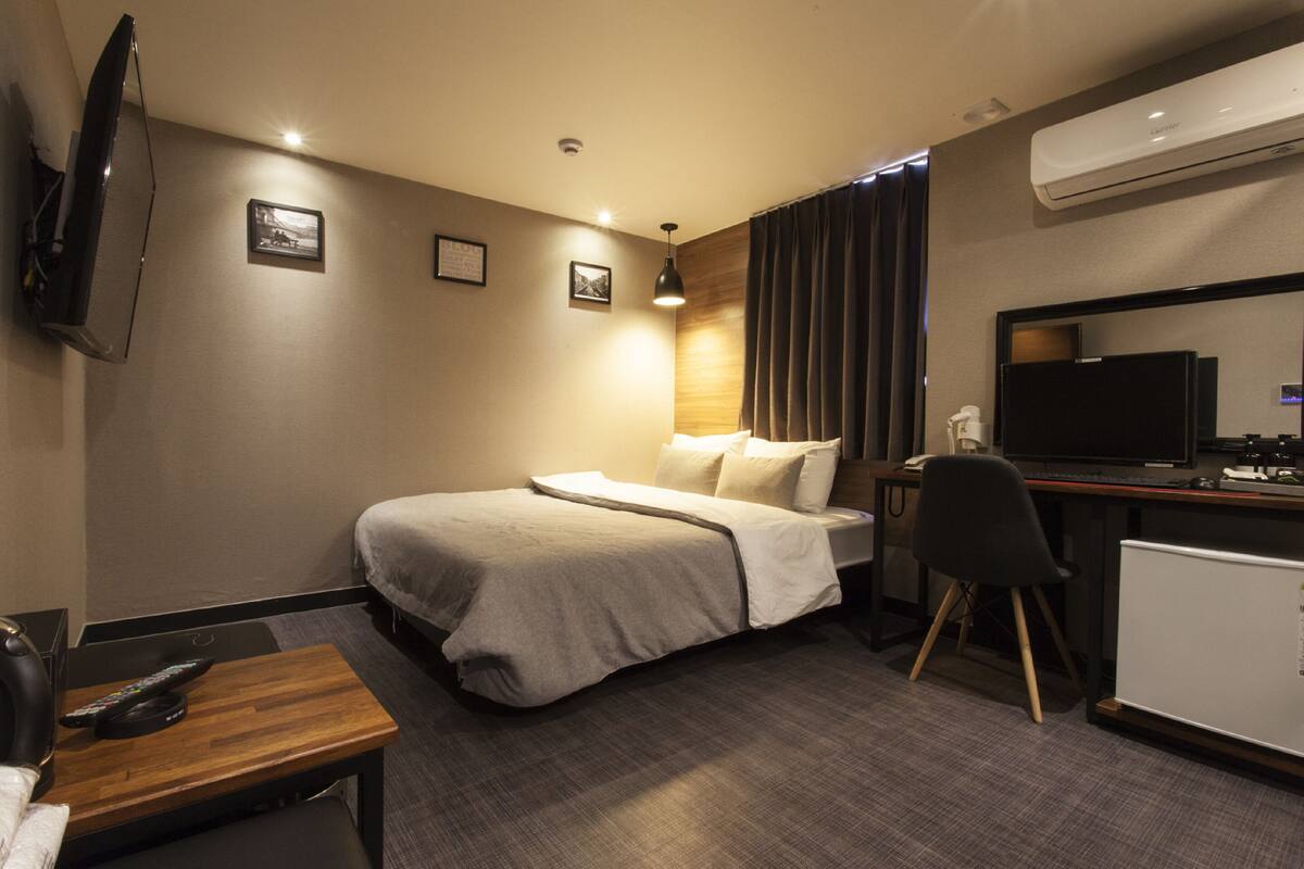 A hotel room with a bed, desk, chair, TV, and air conditioning unit.