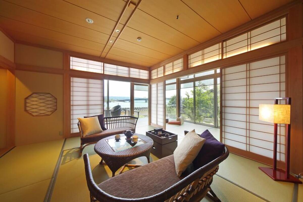 A traditional Japanese-style room with tatami flooring, a low table with cushions, and a view of the sea.
