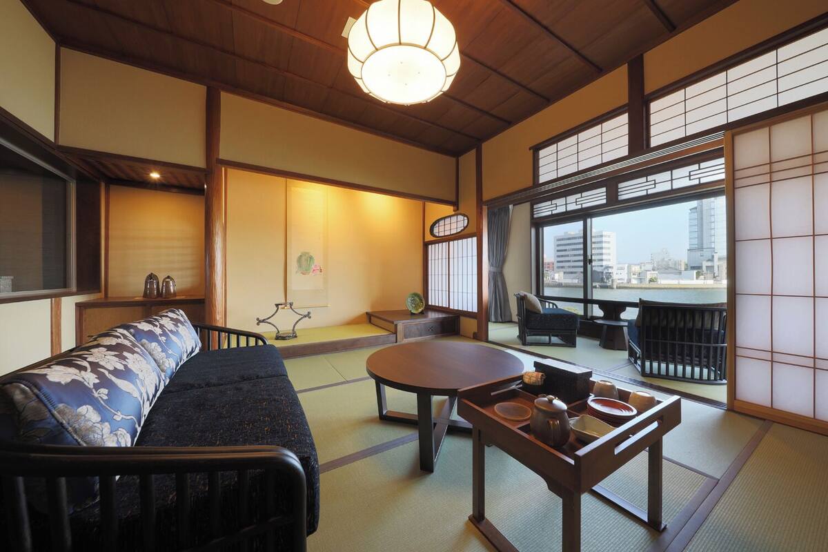 A traditional Japanese room with tatami flooring, a low table with tea set, a sofa, and a view of the city through large windows.