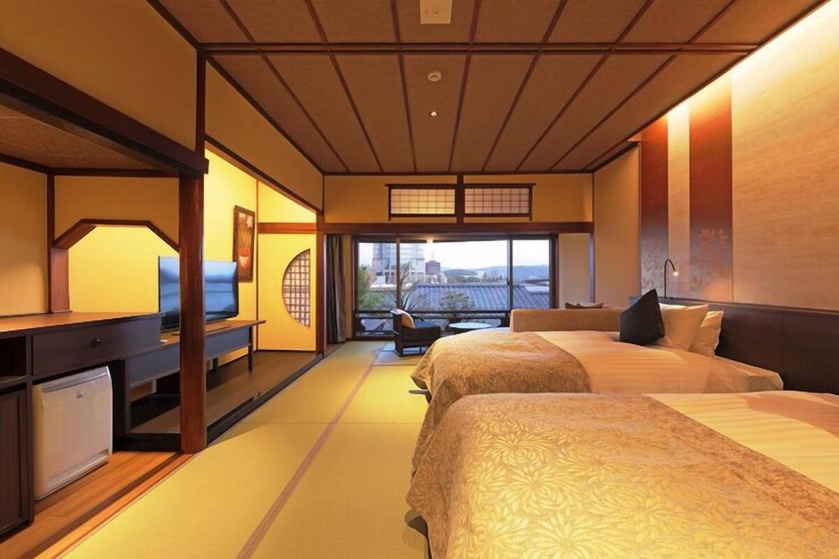 A hotel room with tatami flooring, a traditional Japanese-style bed, a television, and a view of the cityscape.