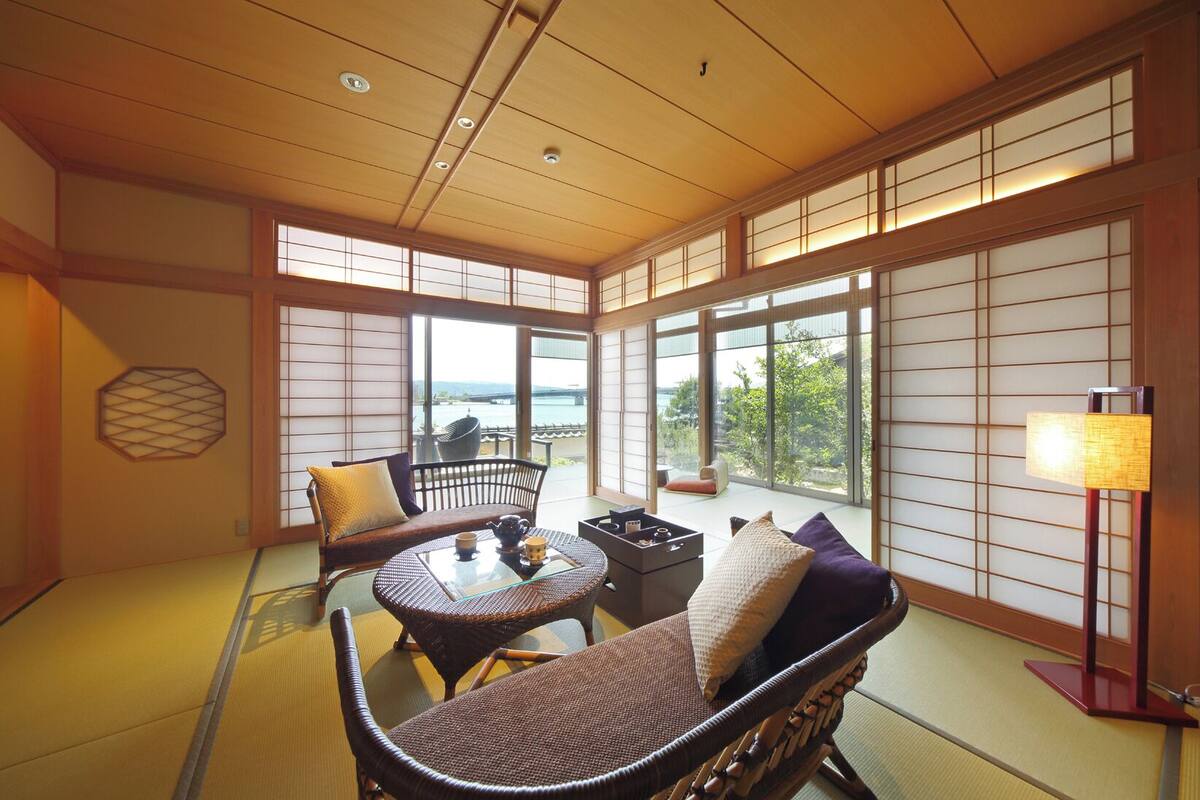 A traditional Japanese room with tatami flooring, a low table with tea set, and a view of the sea through large sliding doors.