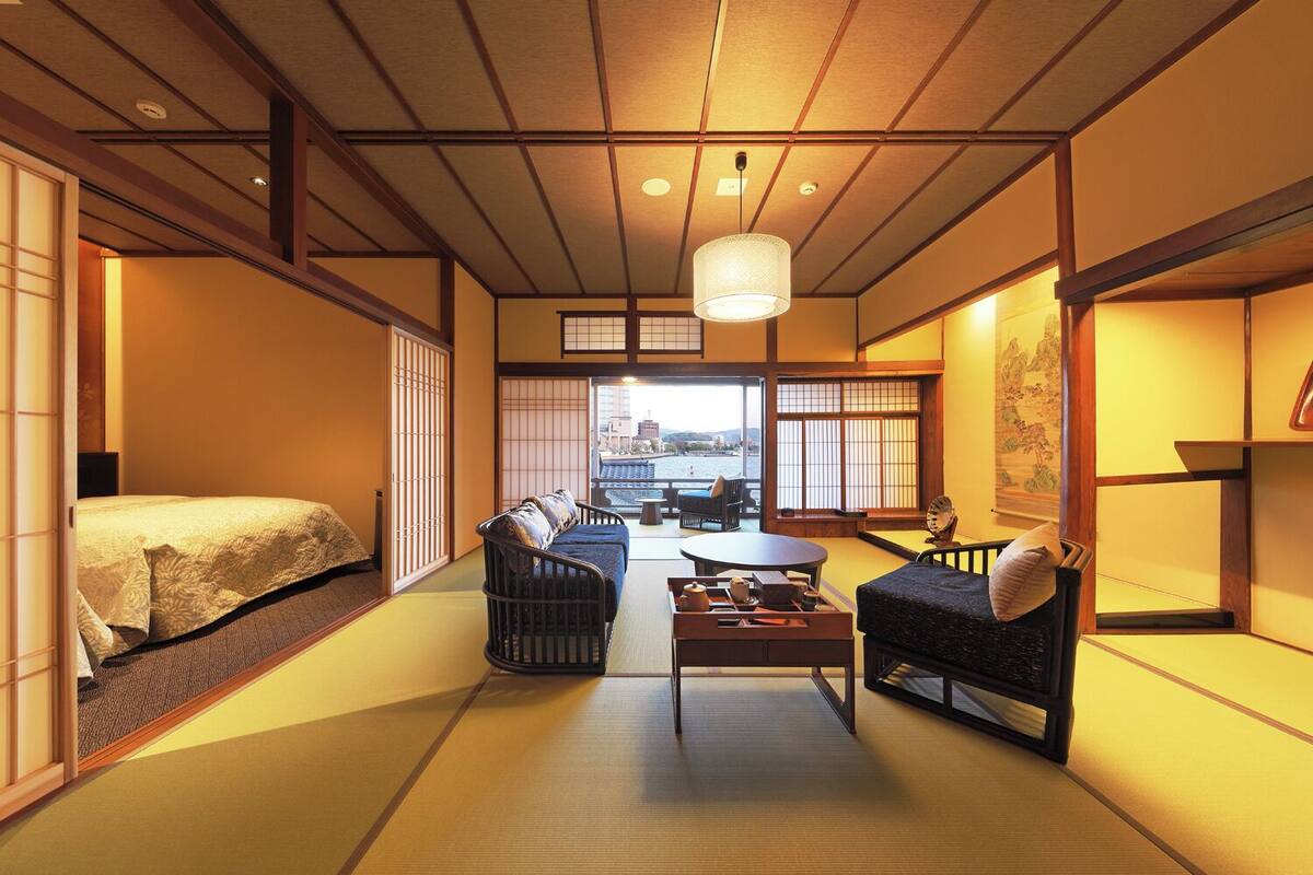 A traditional Japanese-style room with tatami flooring, a low table, two sofas, and a view of a cityscape through large windows.