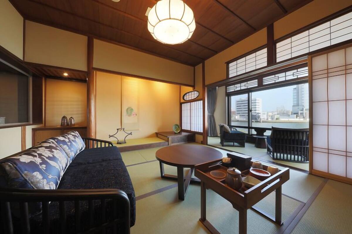A traditional Japanese room with tatami flooring, a low table with tea utensils, a cushioned seating area, and a large window offering a city view.