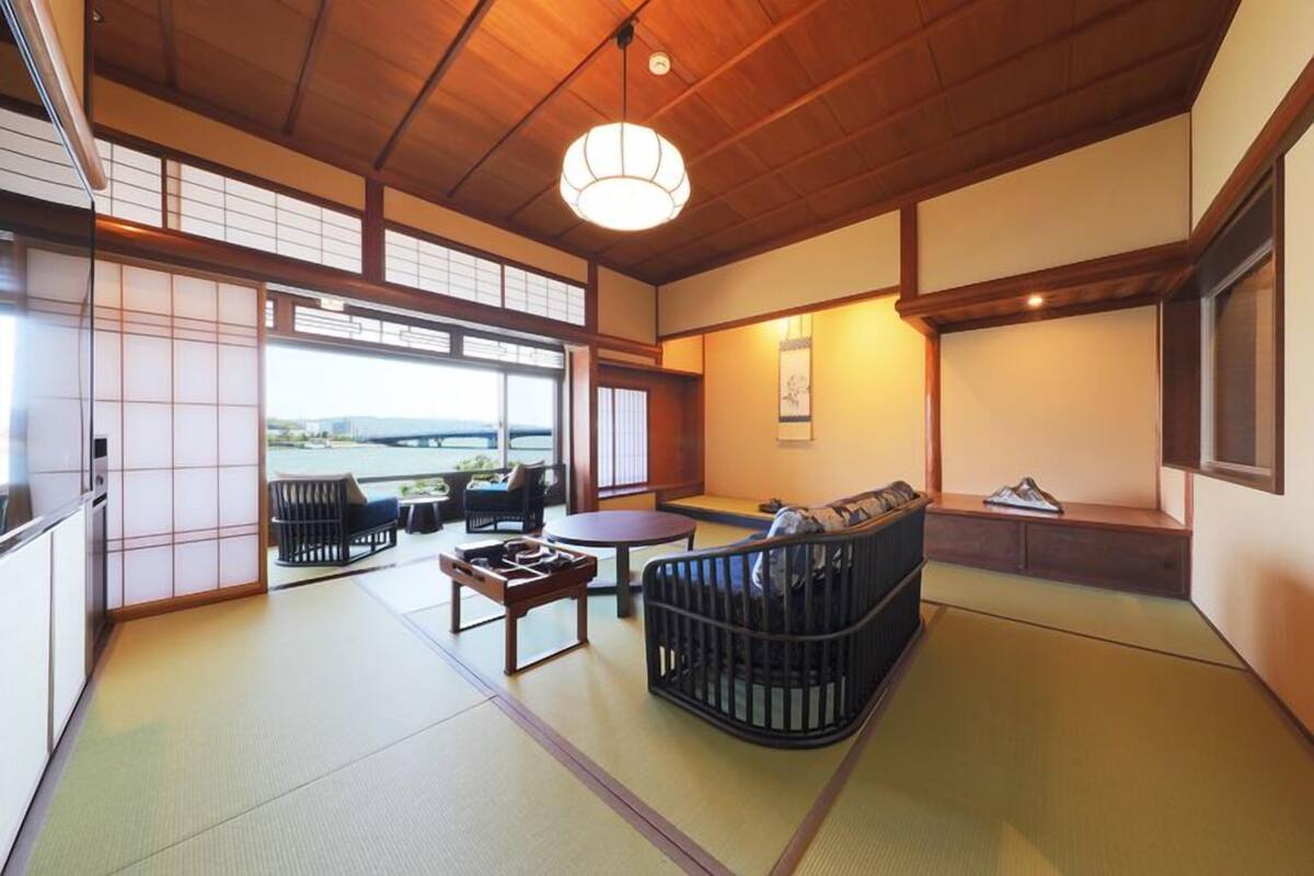 A traditional Japanese room with tatami flooring, a low table, a cushioned seating area, and a view through a sliding door.