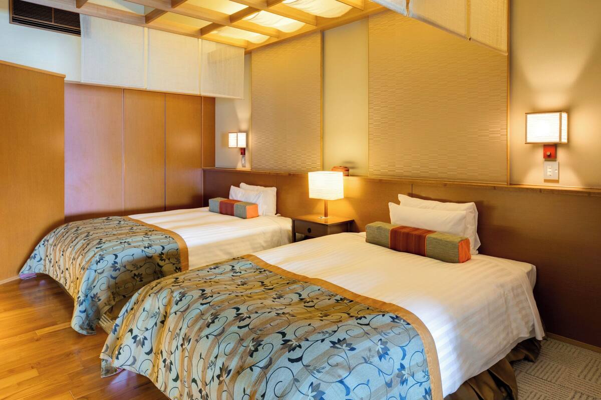 A hotel room with two beds, wooden flooring, a large mirror, and wall-mounted lamps.