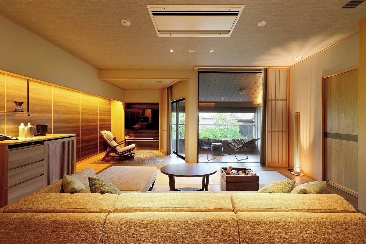 A modern living room with a sofa, a coffee table, and a chair. There is a kitchen area to the left and a balcony with a view.