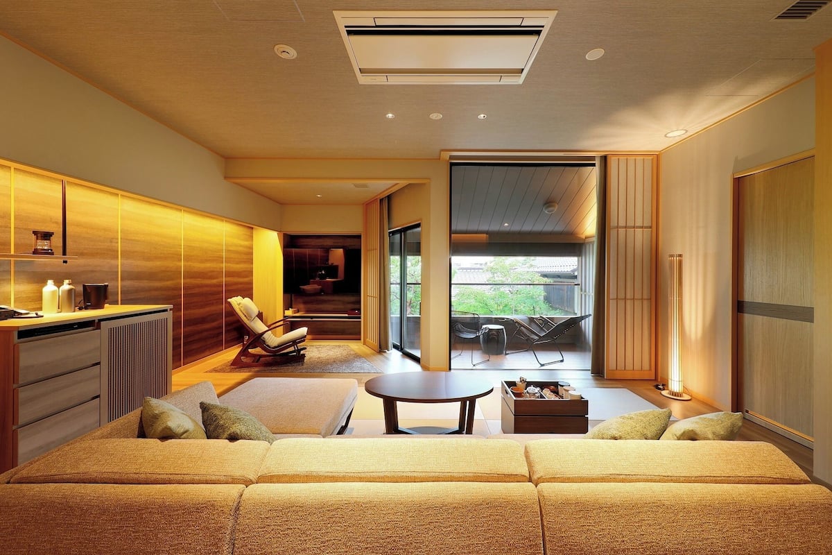 A modern living room with a sofa, a coffee table, and a chair. There is a kitchen area to the left and a balcony with a view.