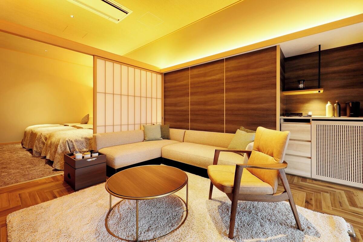 A modern hotel room with a sofa, armchairs, a small table, a cabinet, and a sliding door.