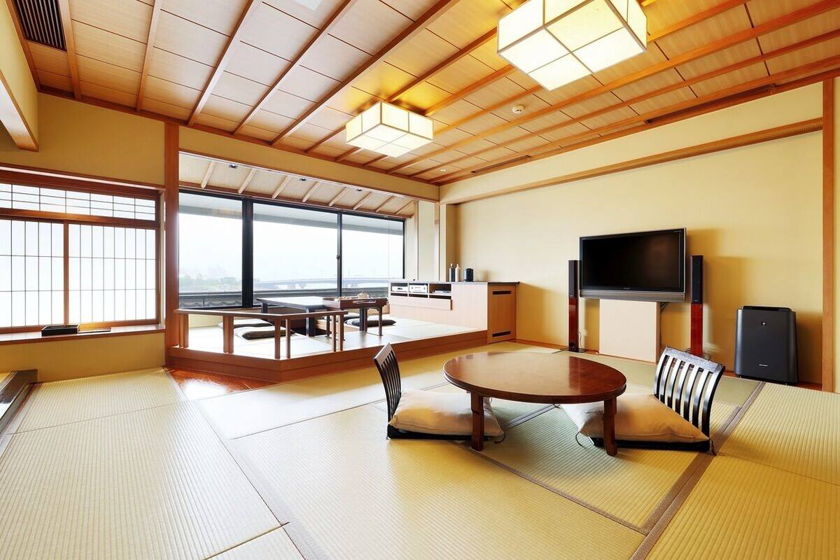A traditional Japanese-style room with tatami flooring, a tatami mat table, a television, and a view through large windows.