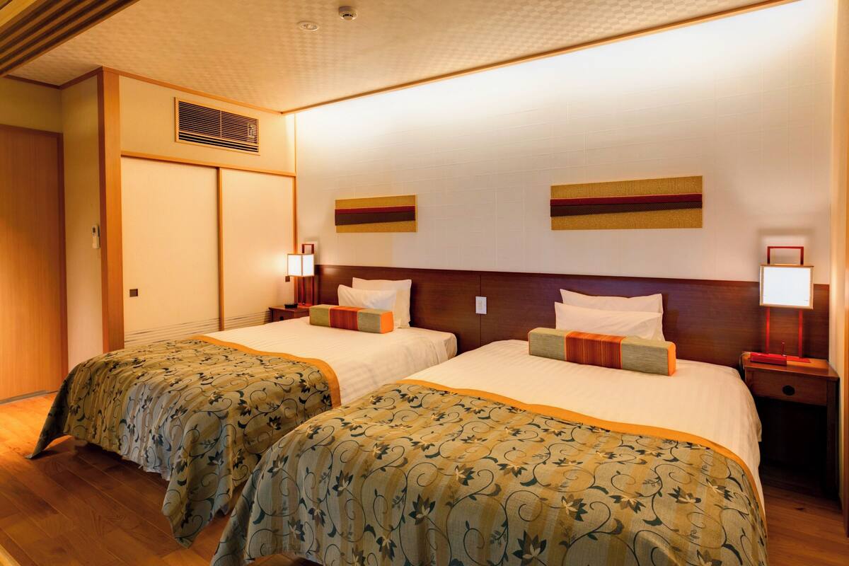 A hotel room with two beds, wooden flooring, a wooden headboard, a wall-mounted vent, and two bedside tables with lamps.