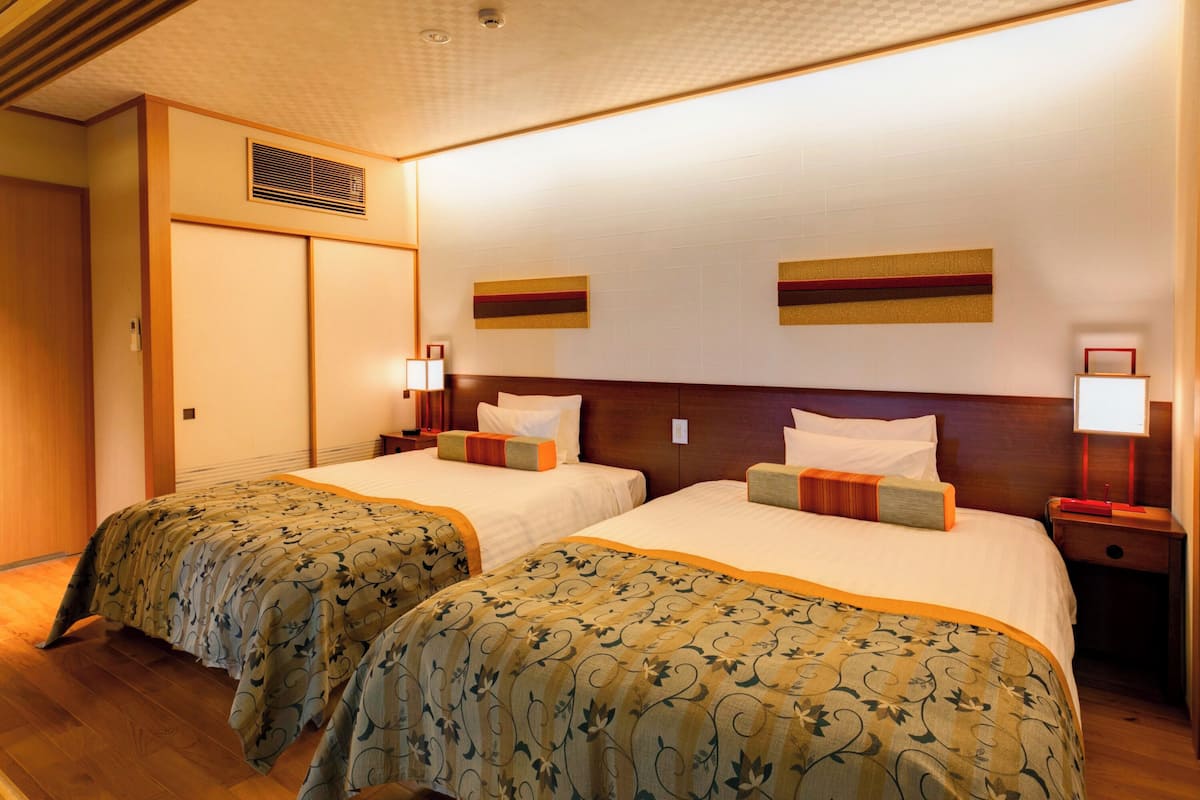 A hotel room with two beds, wooden flooring, a wooden headboard, a wall-mounted vent, and two bedside tables with lamps.