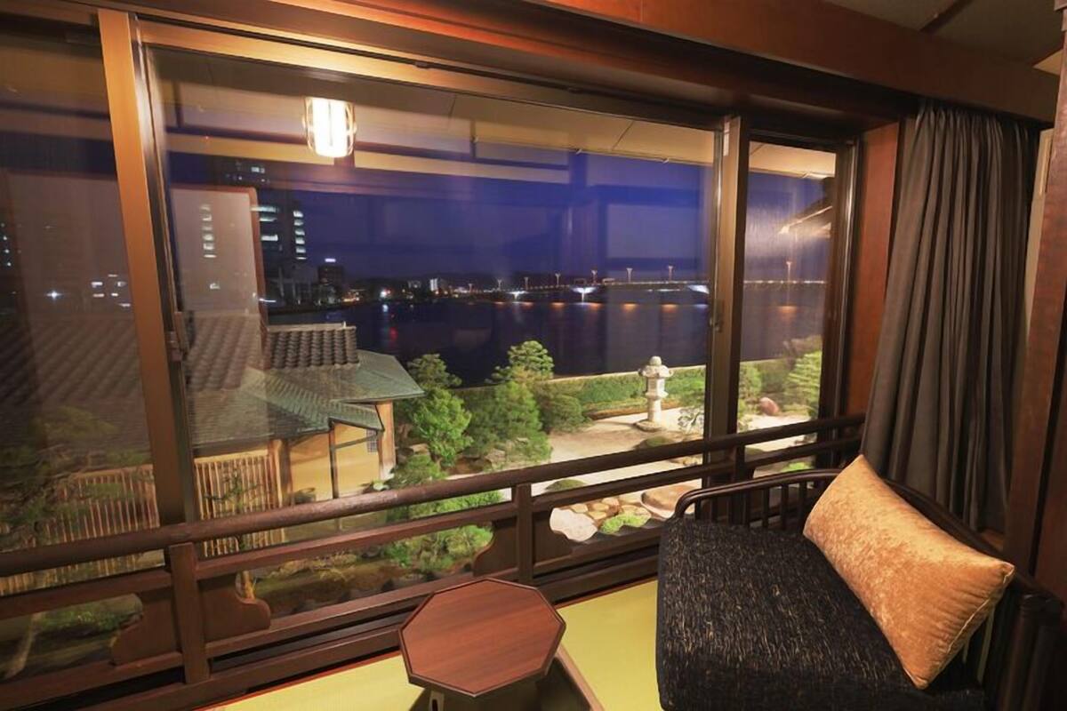 A room with a large window overlooking a body of water and a cityscape at night.