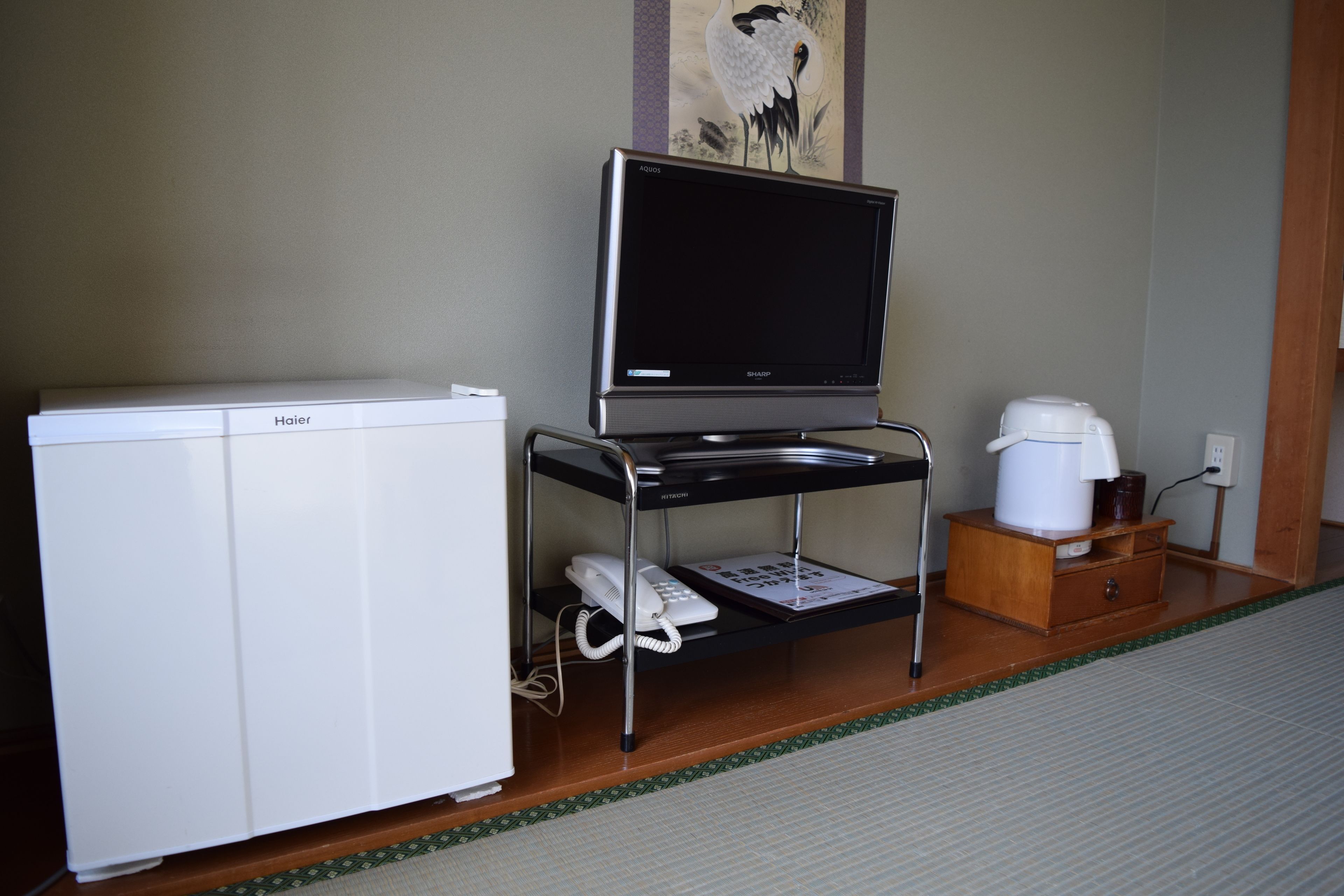 Una habitación con un refrigerador blanco, un televisor de pantalla plana sobre un mueble negro, un gabinete de madera con una cafetera y un teléfono.