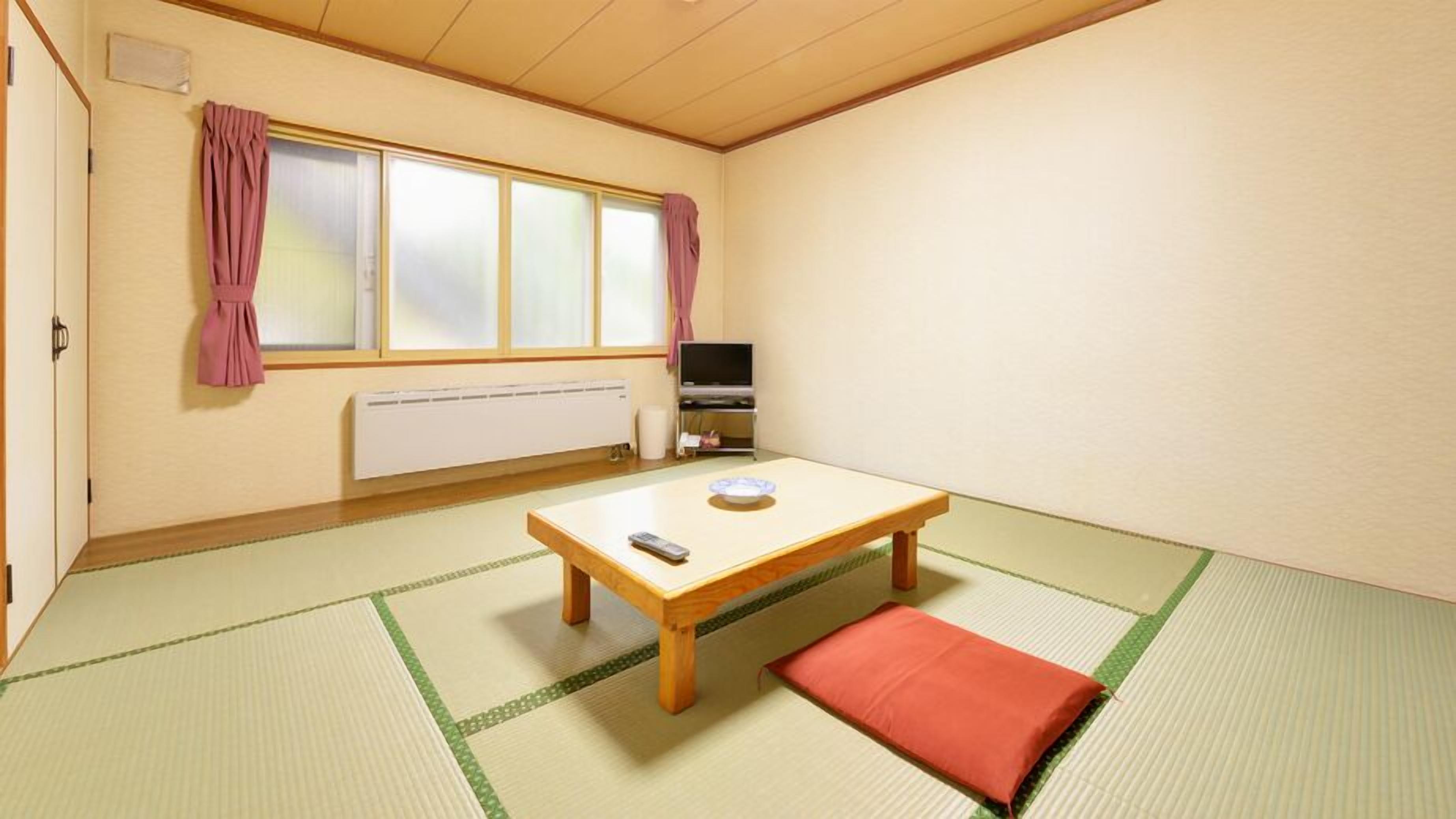 Una stanza con pavimento in tatami, un tavolino in legno, un cuscino rosso, un televisore e una finestra con tende.