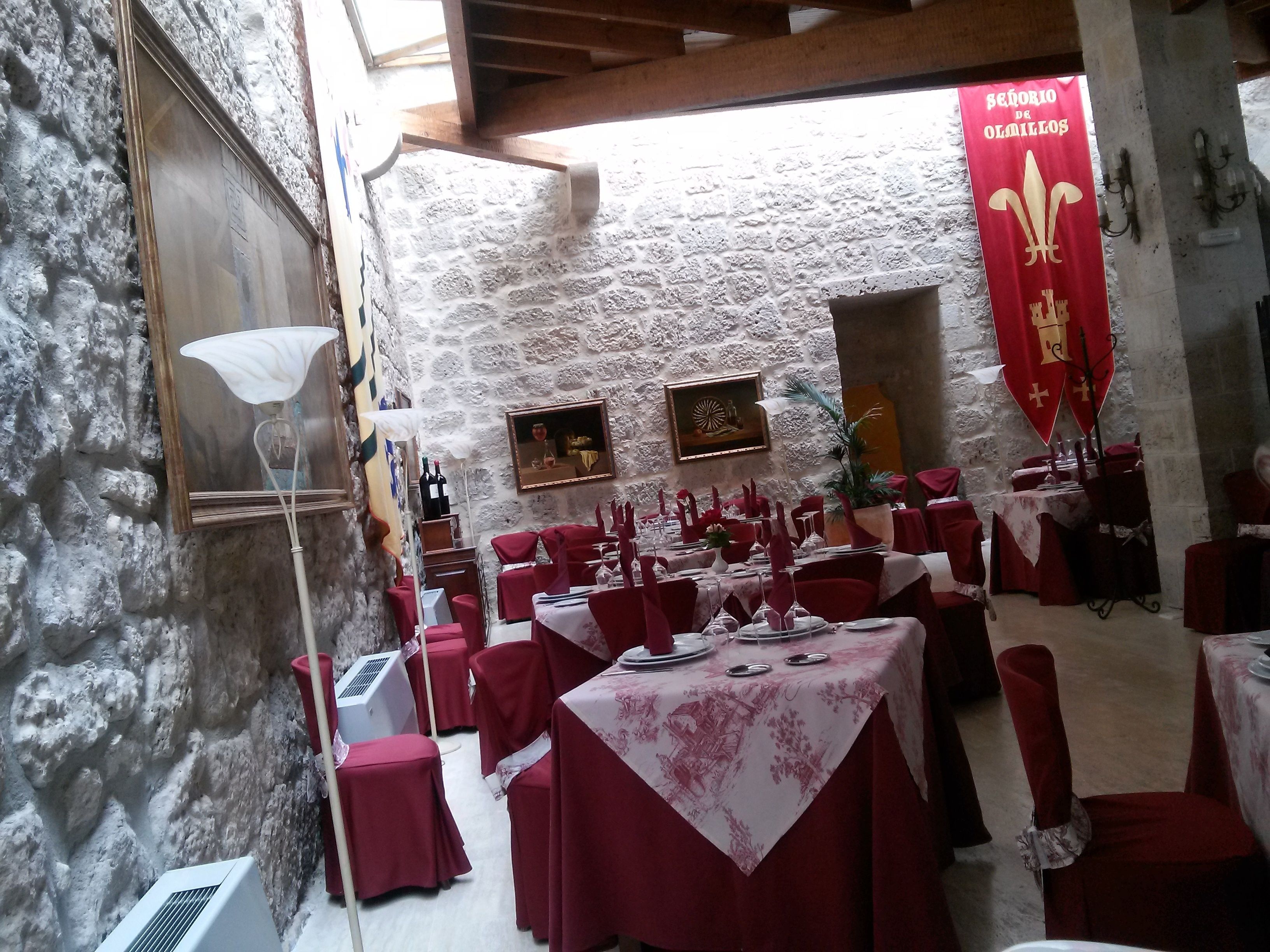 Un comedor con mesas dispuestas para una comida, paredes de piedra y un estandarte con un escudo de armas.