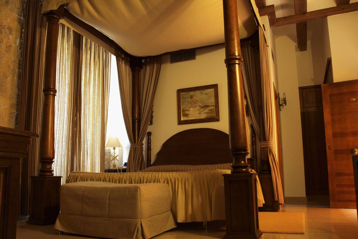 A bedroom with a four-poster bed, a painting on the wall, and a wooden door.