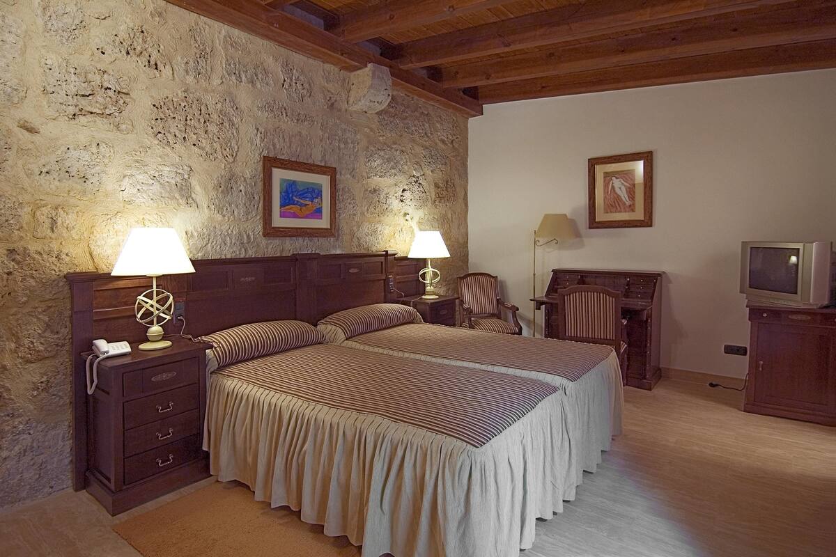 A bedroom with a stone wall, wooden ceiling, two beds with striped bedding, bedside lamps, a chair, a desk with a lamp, and a television.