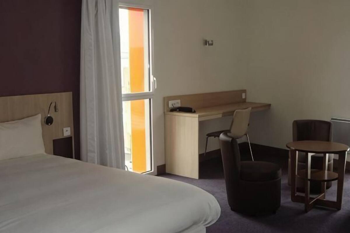 A hotel room with a bed, a desk, a chair, a small table, and a window with curtains.