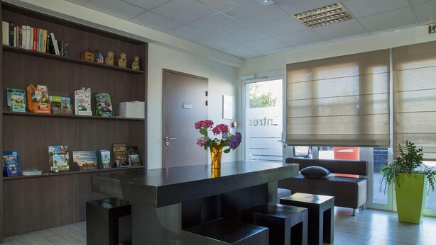 A modern interior with a reception desk, a bookshelf with books and small items, a door, and a window with blinds. - The Originals Access, Hôtel Cholet Gare (Cholet)