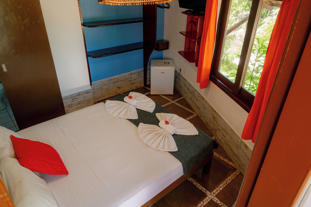 A hotel room with a bed, a small refrigerator, a window with curtains, and a shelf with a television.