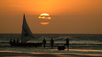 Um pôr do sol sobre o oceano, com um veleiro e pessoas na praia.