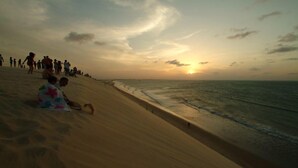 Escena de playa con gente caminando y sentada sobre la arena, un atardecer al fondo y el océano a la derecha.