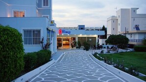 A hotel entrance with a tiled pathway leading to the front door, surrounded by greenery and adjacent buildings. - Miramare Bay Hotel (Karpathos)