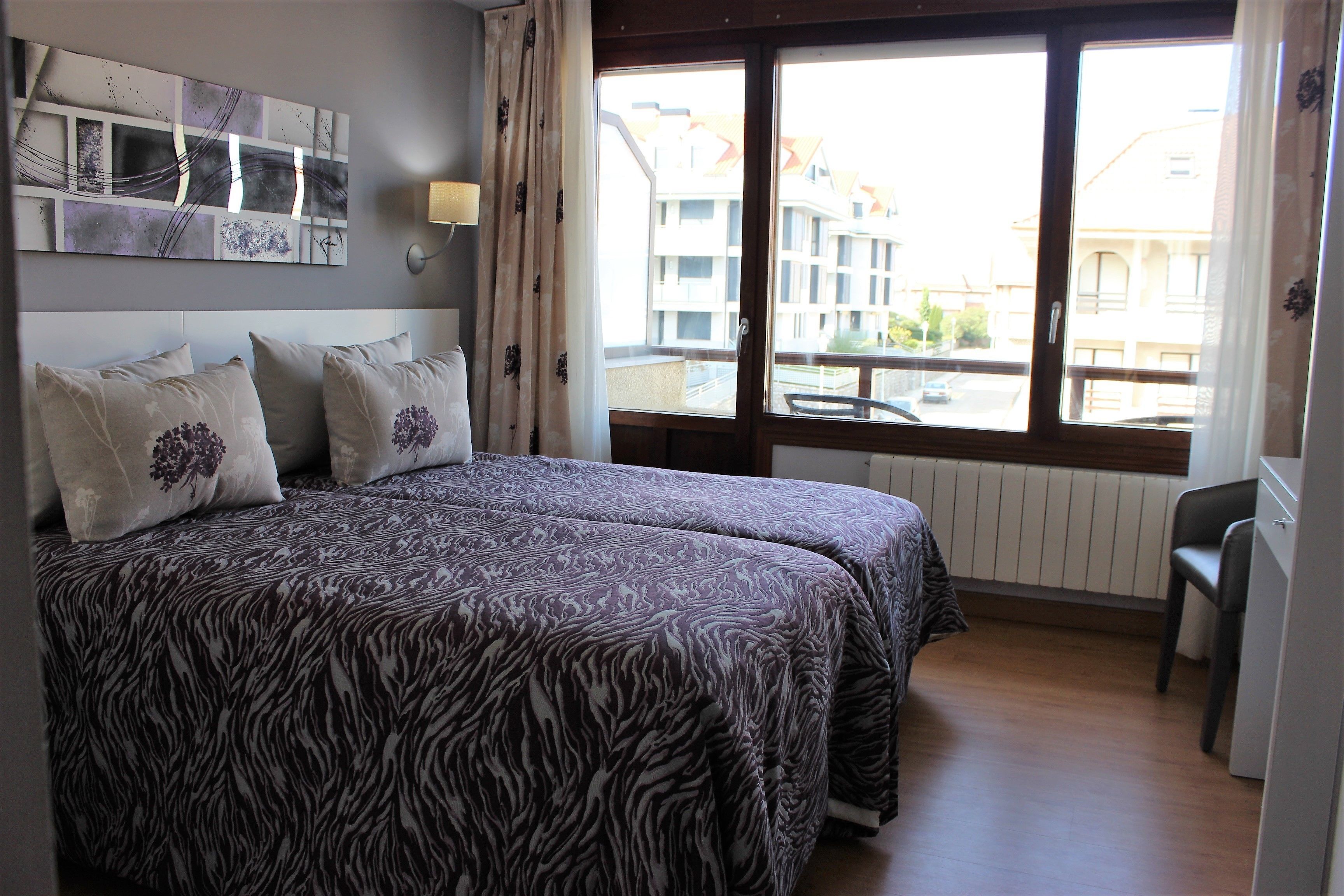 A bedroom with a bed, a window with a view of buildings, and a wall-mounted lamp.