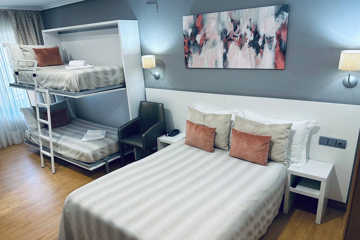 A hotel room with a bunk bed, a desk, a chair, a bedside table with a lamp, and a painting on the wall.