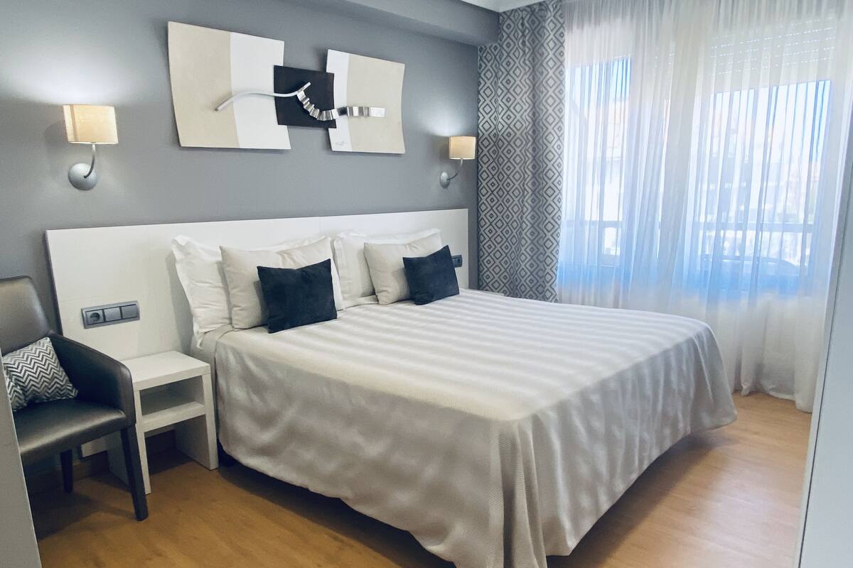 A modern hotel room with a large bed, a bedside table, a chair, and a wall-mounted light fixture.