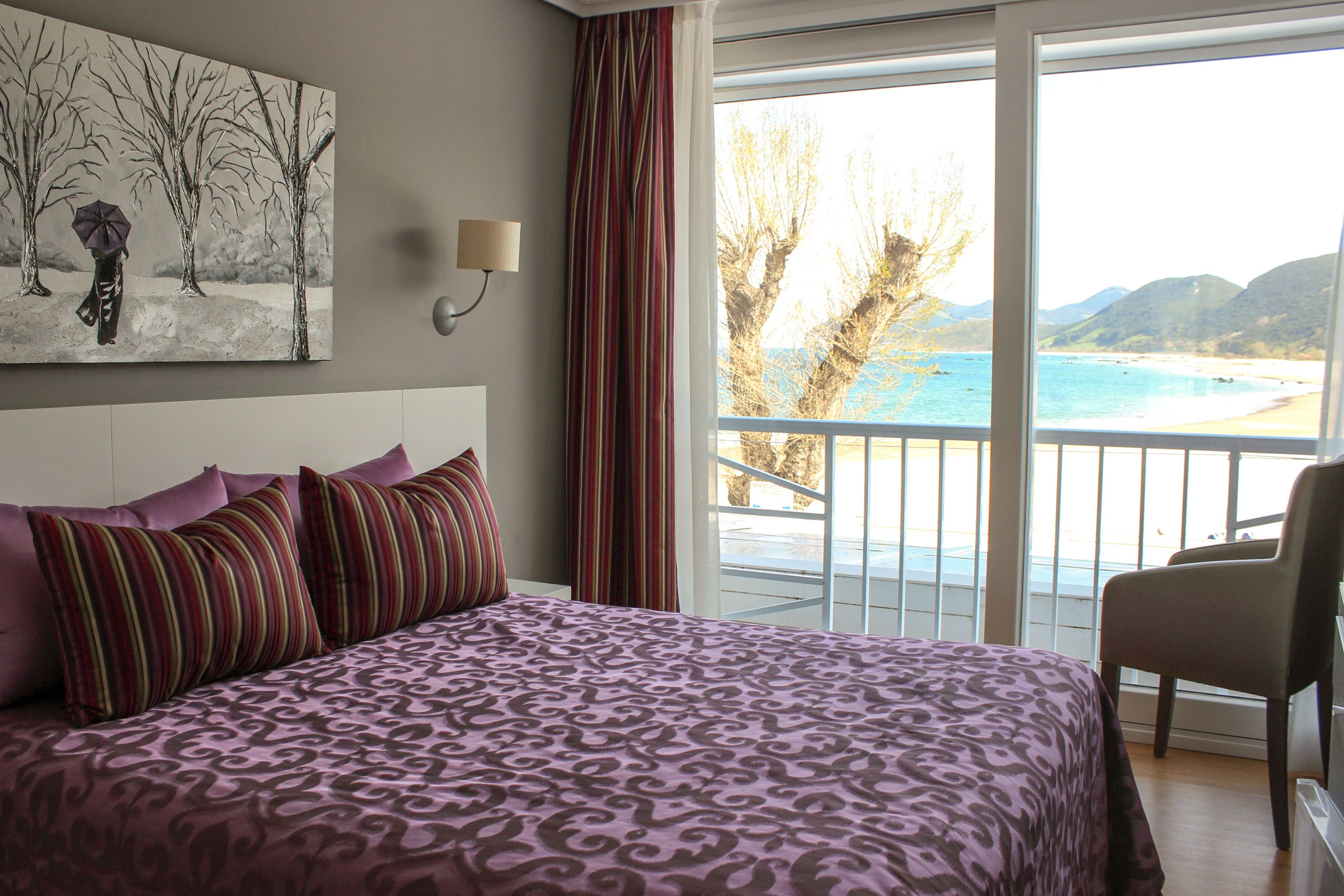 Un dormitorio con una cama grande, un cuadro de una persona con un paraguas, una ventana con vista a una playa y montañas, y una silla junto a la ventana.