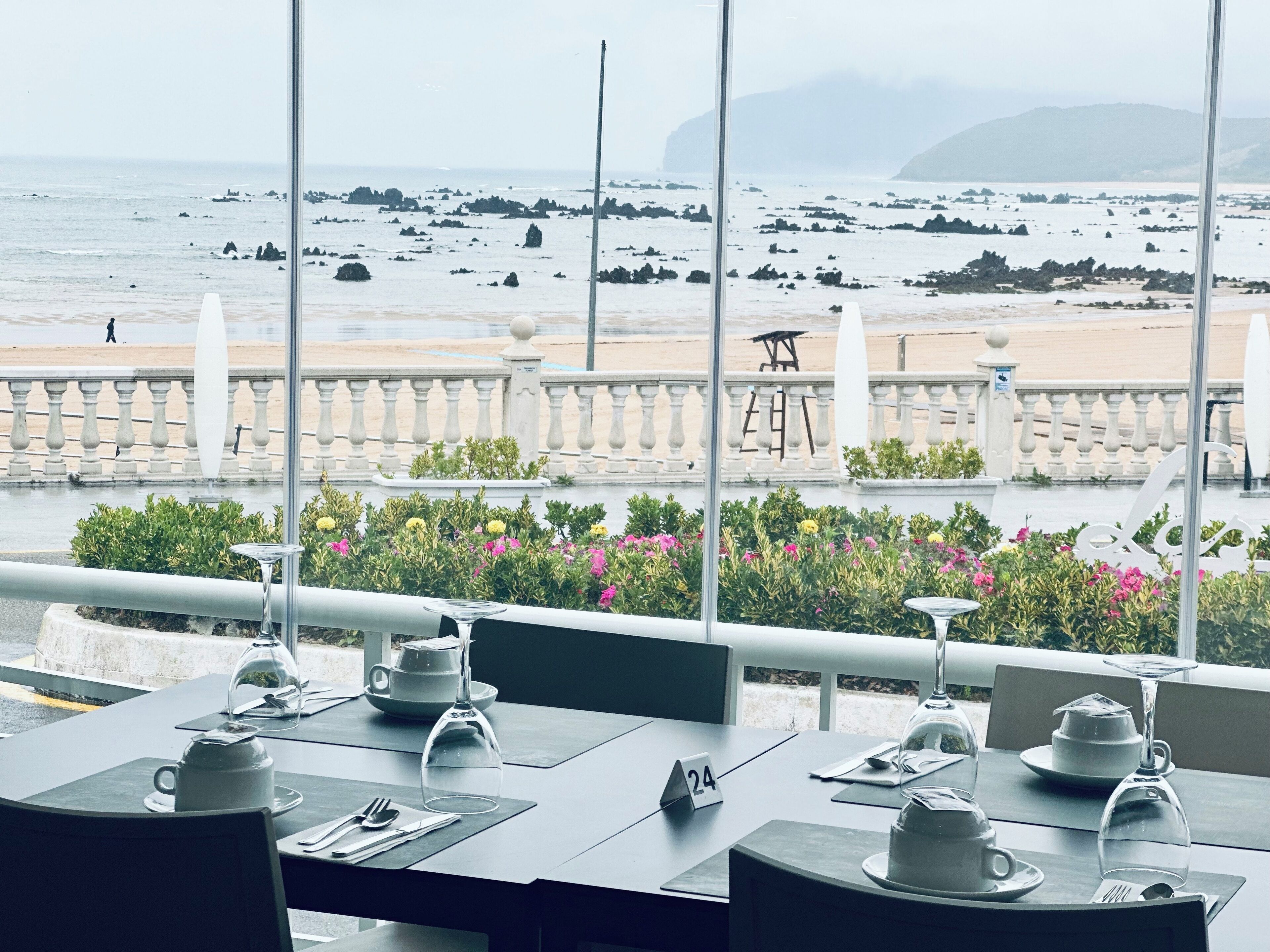 A restaurant with a view of the beach and rocky coastline. Tables are set with white tableware and a wine glass.