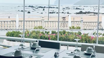 A restaurant with a view of the beach and rocky coastline. Tables are set with white tableware and a wine glass.
