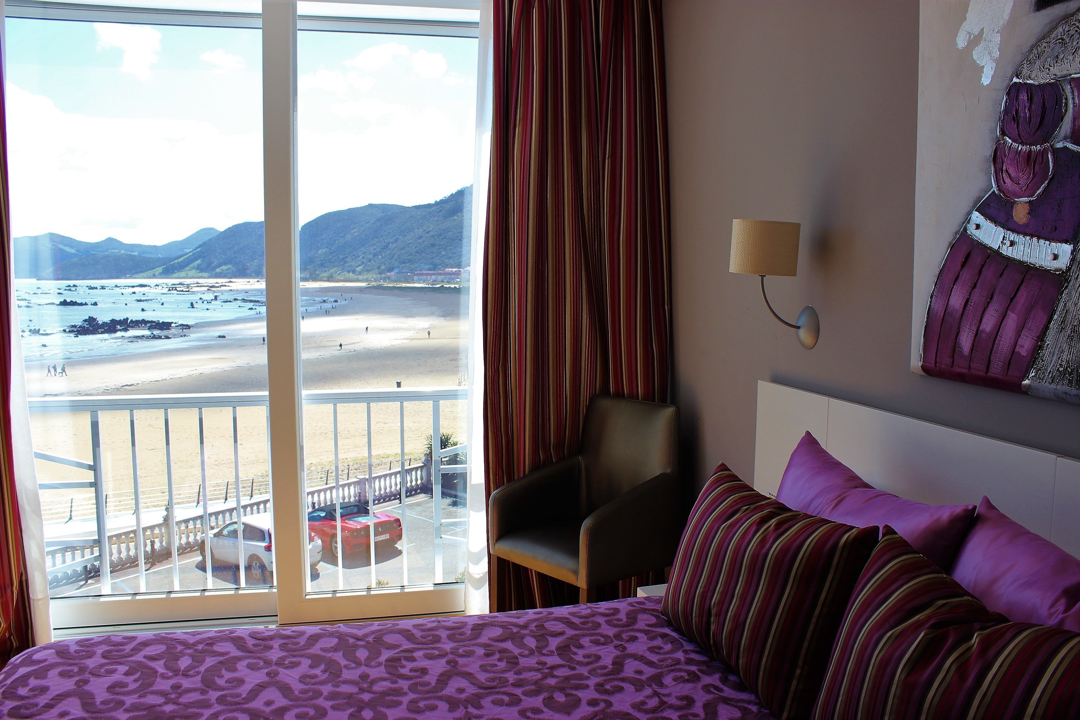 Une chambre d’hôtel avec un lit, une chaise, une fenêtre donnant sur une plage et des montagnes, et une lampe fixée au mur.